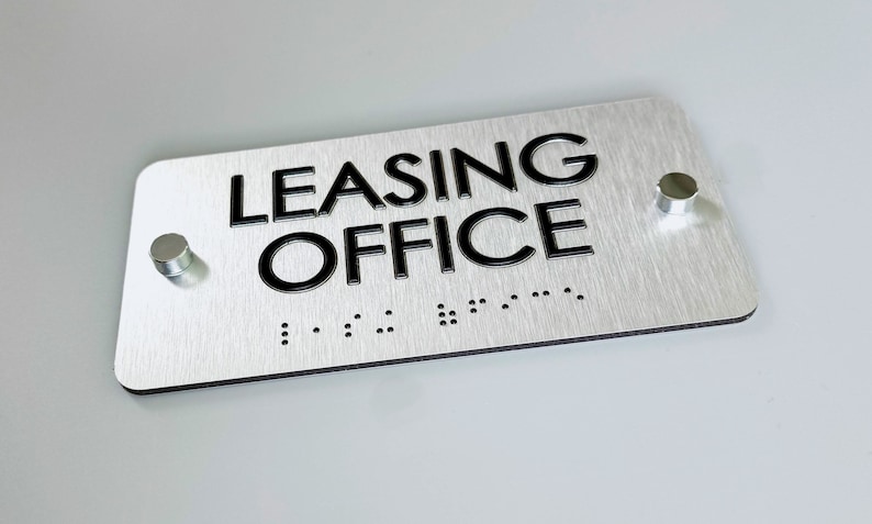 K&ouml;nnte beinhalten: Ein silbernes Metallschild mit schwarzer Beschriftung, auf dem "LEASING OFFICE" steht, mit Braille unter dem Text. Das Schild hat zwei silberne Montagepfosten.