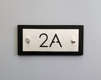 Könnte beinhalten: Ein schwarzes und silbernes Hausnummernschild mit der Nummer "2A" in Schwarz auf silbernem Hintergrund. Das Schild hat zwei silberne Schrauben an jeder Seite.