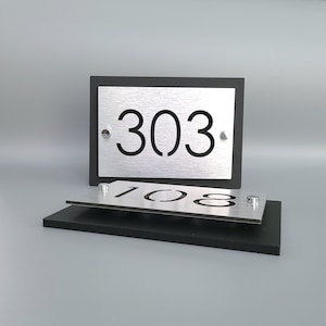 May include: Two silver house numbers with black lettering on a black background. The top number is 303 and the bottom number is 108.