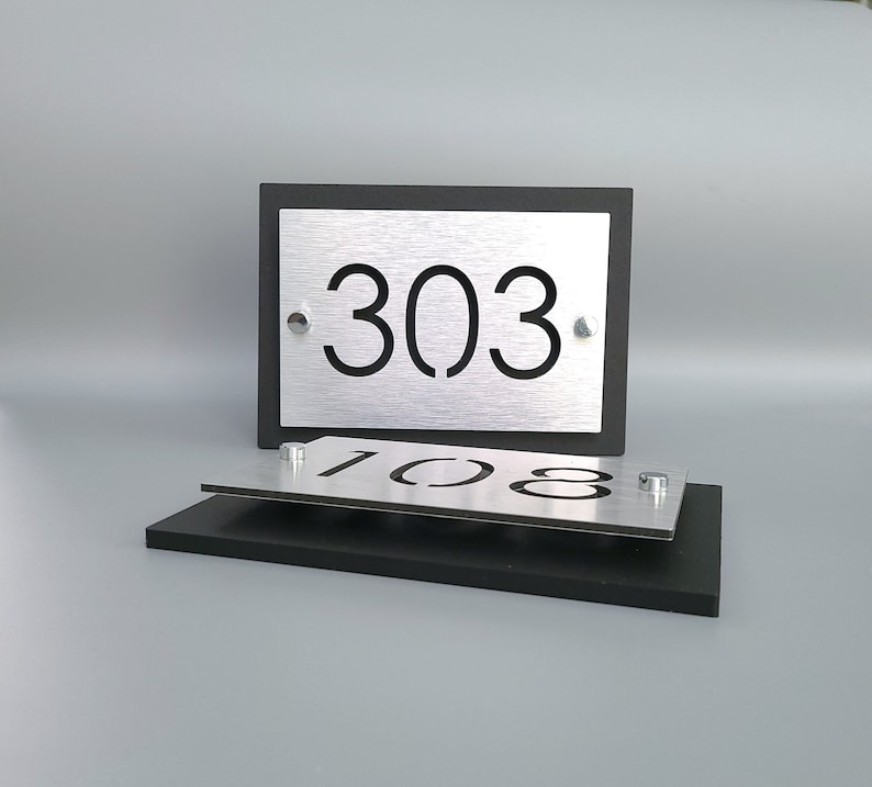 May include: Two silver house numbers with black lettering on a black background. The top number is 303 and the bottom number is 108.