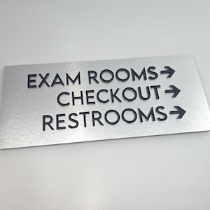 May include: A rectangular brushed silver metal sign featuring raised black lettering and directional arrows. The text reads EXAM ROOMS, CHECKOUT, and RESTROOMS, each followed by a right-pointing arrow, providing clear navigation for a facility.