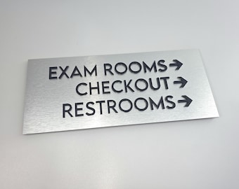 May include: A rectangular brushed silver metal sign featuring raised black lettering and directional arrows. The text reads EXAM ROOMS, CHECKOUT, and RESTROOMS, each followed by a right-pointing arrow, providing clear navigation for a facility.