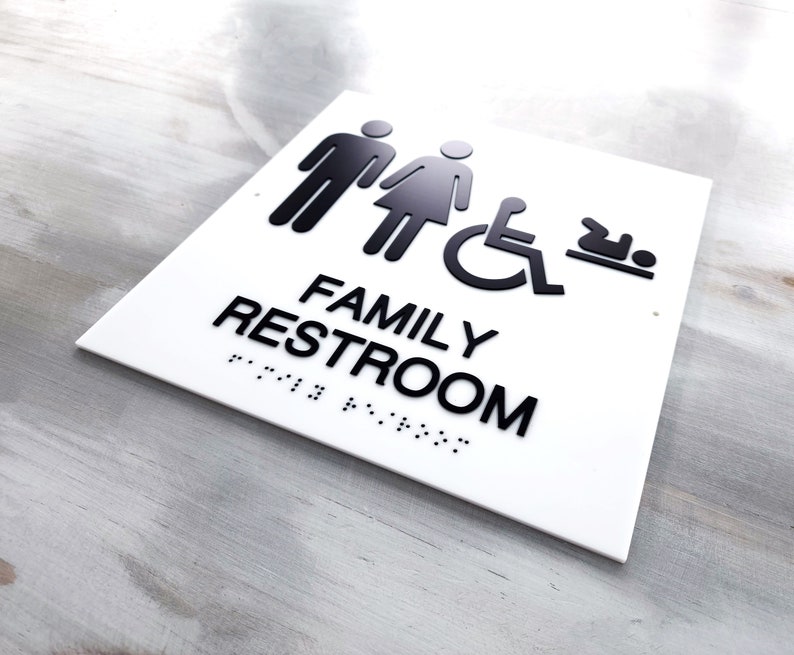 Pu&ograve; includere: Un cartello bianco con testo nero e icone che recita "FAMILY RESTROOM" con braille sotto. Le icone rappresentano un uomo, una donna, una persona su sedia a rotelle e un bambino.