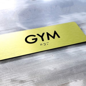 May include: A rectangular, gold-colored sign with the word "GYM" in black, sans-serif font. Below the word "GYM" is braille. The sign is set against a light gray and white wooden background.