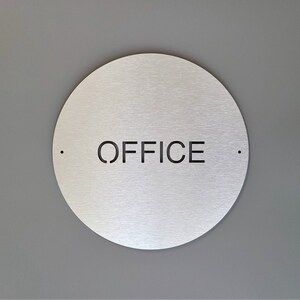 May include: A circular, brushed metal sign with the word "OFFICE" cut out in black. The sign is mounted on a grey wall. Small black dots are either side of the word.
