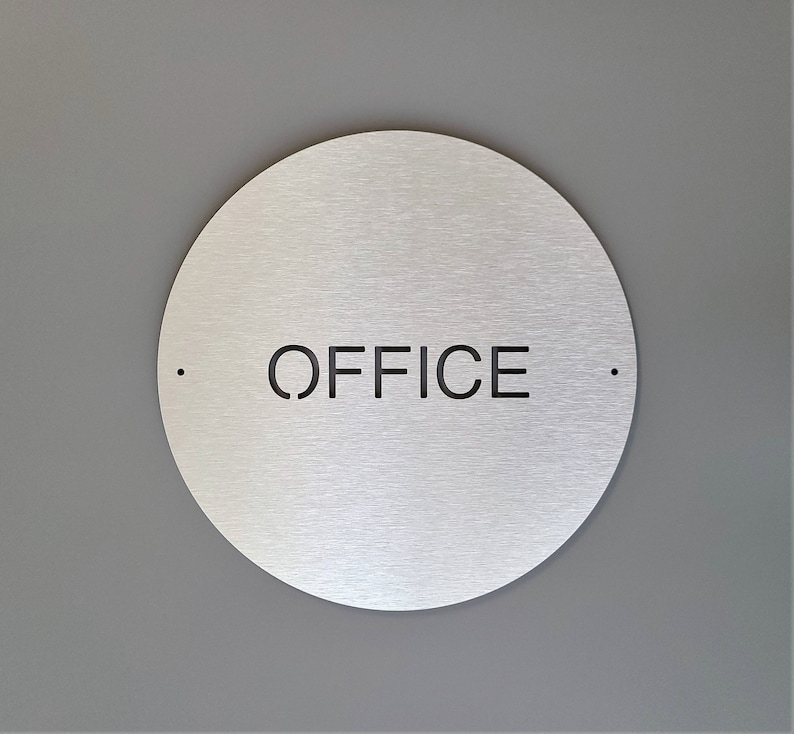 May include: A circular, brushed metal sign with the word "OFFICE" cut out in black. The sign is mounted on a grey wall. Small black dots are either side of the word.