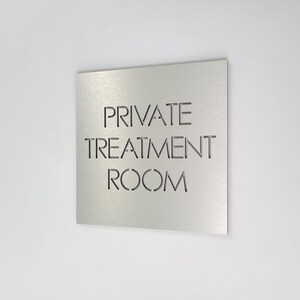 May include: A square, brushed metal sign with the words "PRIVATE TREATMENT ROOM" cut out in a sans-serif font. The sign is mounted on a white wall, angled slightly to the left.