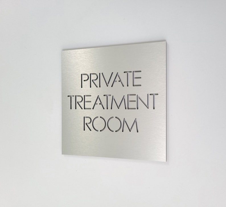 May include: A square, brushed metal sign with the words "PRIVATE TREATMENT ROOM" cut out in a sans-serif font. The sign is mounted on a white wall, angled slightly to the left.