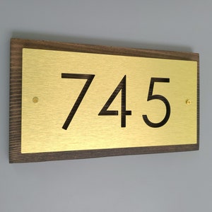 May include: A gold metal house number plaque with the number 745 on it. The plaque is mounted on a dark brown wooden frame.
