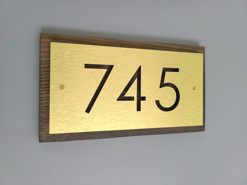 May include: A gold metal house number plaque with the number 745 on it. The plaque is mounted on a dark brown wooden frame.