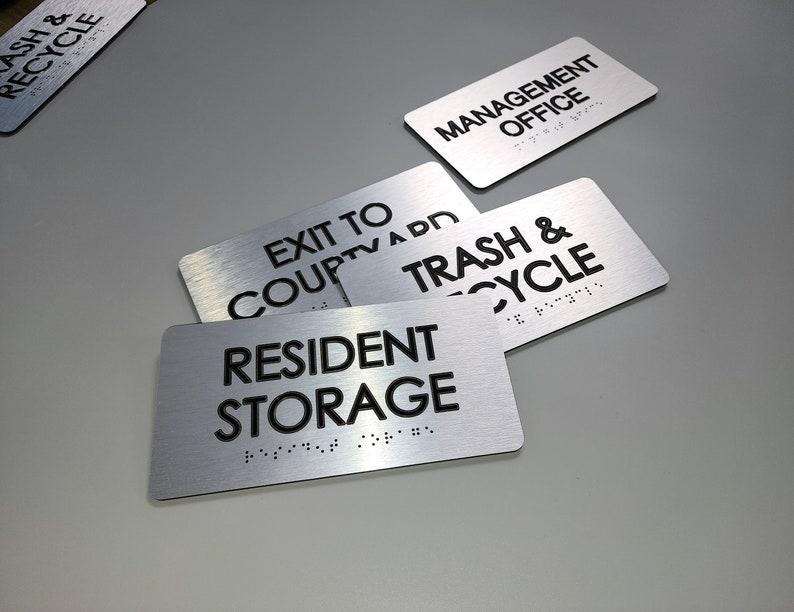 K&ouml;nnte beinhalten: Vier silberne Metallschildchen mit schwarzem Text und Braille-Punkten. Die Schilder zeigen "Trash & Recycle", "Resident Storage", "Exit to Couptva App" und "Management Office".