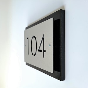 May include: A black and silver house number plaque with the number 104 cut out. The plaque is mounted on a white wall.