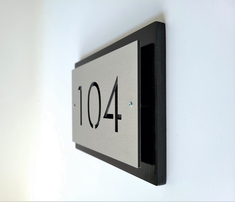 May include: A black and silver house number plaque with the number 104 cut out. The plaque is mounted on a white wall.