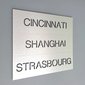 May include: A silver metal sign with three lines of text. The top line reads "CINCINNATI", the middle line reads "SHANGHAI", and the bottom line reads "STRASBOURG".