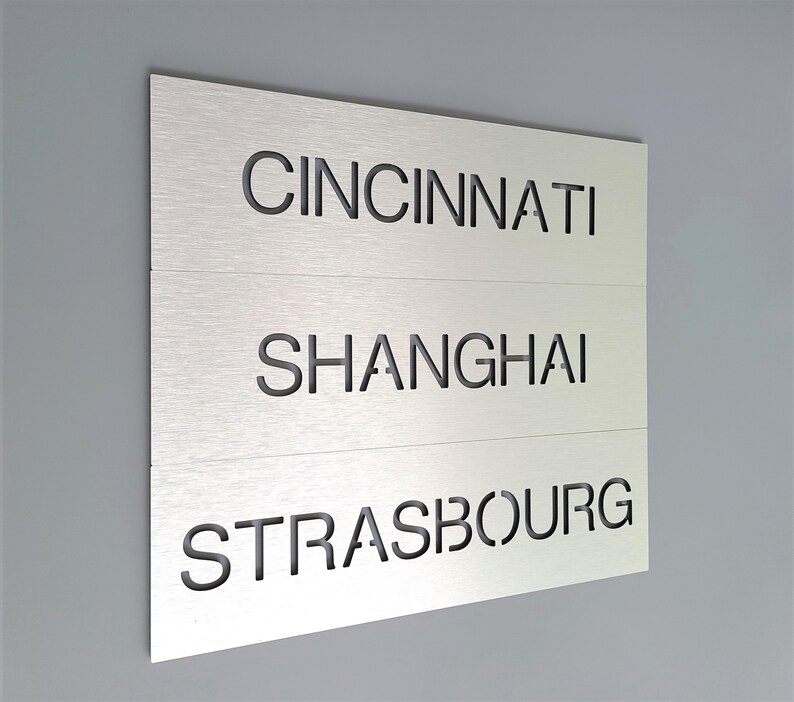 May include: A silver metal sign with three lines of text. The top line reads "CINCINNATI", the middle line reads "SHANGHAI", and the bottom line reads "STRASBOURG".