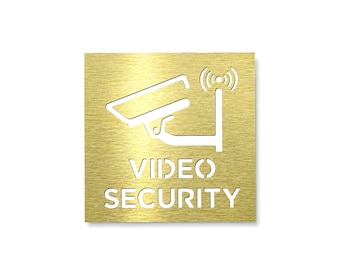 Könnte beinhalten: Ein quadratisches Schild aus goldfarbenem Metall mit gebürsteter Oberfläche, das eine ausgeschnittene Grafik einer Überwachungskamera mit einem Funksymbol darüber zeigt. Der Text VIDEO SECURITY ist in Großbuchstaben darunter ausgeschnitten.