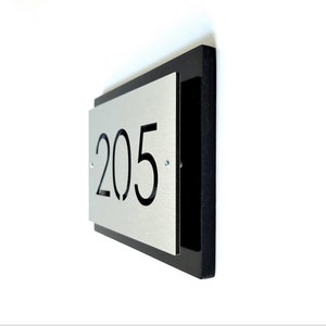 May include: A silver metal house number sign with black numbers "205" on a black background.