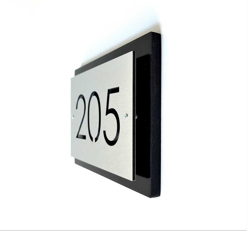 May include: A silver metal house number sign with black numbers "205" on a black background.