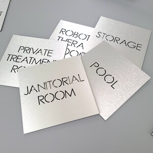 May include: Silver metal square signs with black text. Signs include "Private Treatment Room", "Robot Therapy Room", "Storage", "Pool", and "Janitorial Room". The signs are on a gray surface.