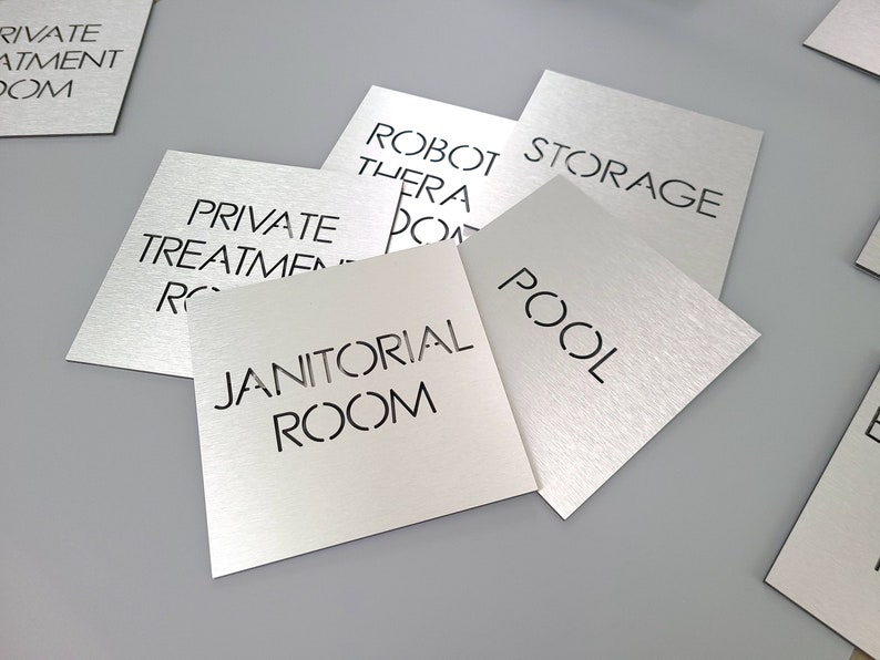 May include: Silver metal square signs with black text. Signs include "Private Treatment Room", "Robot Therapy Room", "Storage", "Pool", and "Janitorial Room". The signs are on a gray surface.