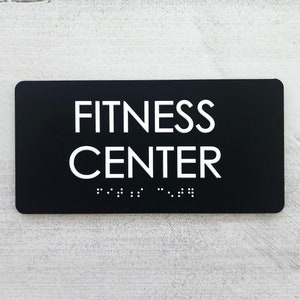 K&ouml;nnte beinhalten: Schwarzes rechteckiges Schild mit wei&szlig;em Text "FITNESS CENTER" und Braille-Schrift darunter.