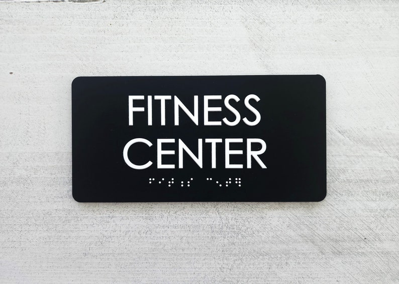 K&ouml;nnte beinhalten: Schwarzes rechteckiges Schild mit wei&szlig;em Text "FITNESS CENTER" und Braille-Schrift darunter.