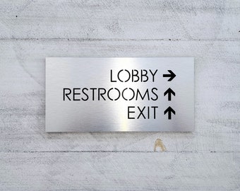 May include: A silver metal sign with black text and arrows indicating directions. The sign reads "Lobby", "Restrooms", and "Exit" with arrows pointing right, up, and up respectively.