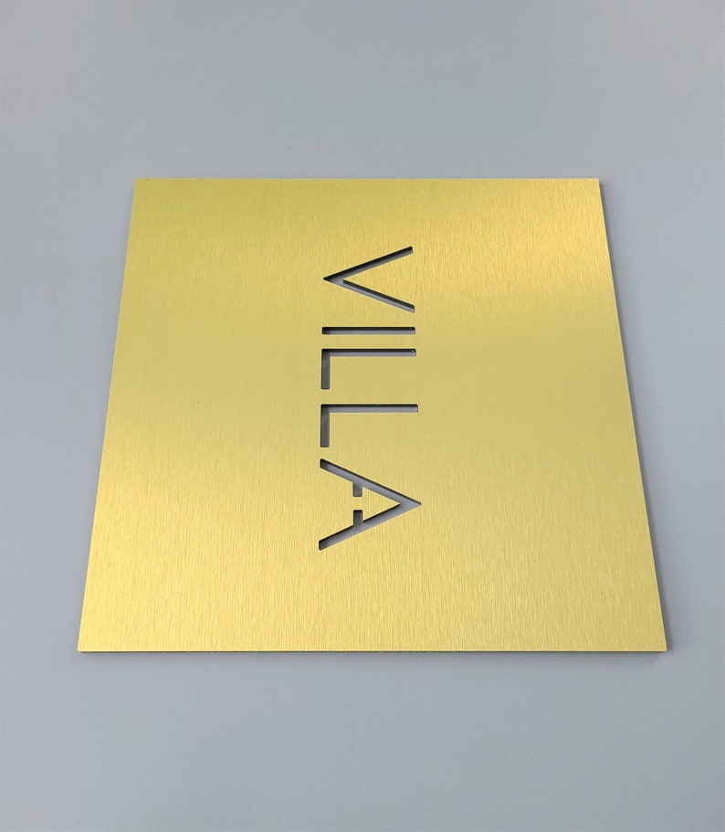 May include: A gold-colored rectangular sign with the word "VILLA" cut out vertically. The sign has a brushed metal texture and is set against a gray background. The sign is likely for a home or business.
