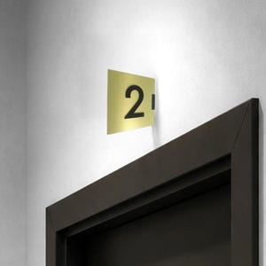 May include: A rectangular gold-coloured metal door sign featuring the black number 2 prominently displayed on its surface. The sign is mounted perpendicularly to the wall, extending outward from the side of a dark brown door frame.