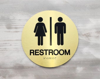 May include: A round gold sign with black silhouettes of a woman and a man, and the word "RESTROOM" in black letters. The sign also has braille lettering.