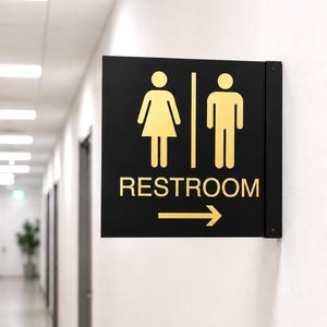 May include: A square black wall-mounted restroom sign featuring gold-colored icons of a person in a dress and a person in pants, separated by a vertical line. The text RESTROOM is printed in gold below the icons, with a gold arrow pointing right.