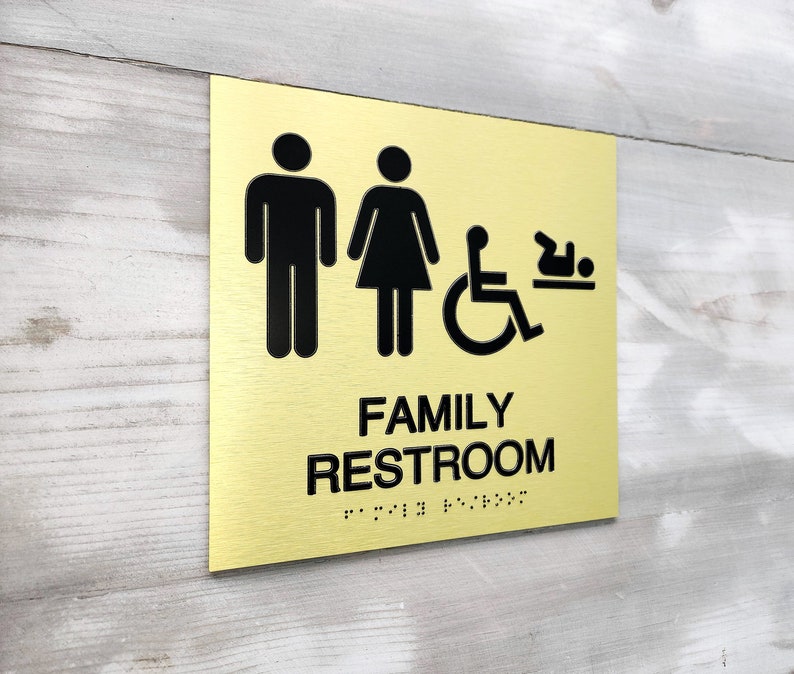 Pu&ograve; includere: Un cartello in metallo dorato con testo nero e icone. Il cartello recita "FAMILY RESTROOM" con il braille sotto. Le icone rappresentano un uomo, una donna, una persona su sedia a rotelle e un bambino.