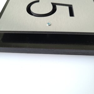 May include: A silver metal house number sign with black framing and the number "305" cut out. The sign is mounted with a screw.