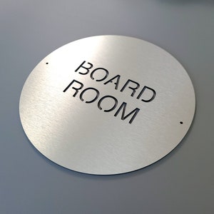 May include: A circular sign made of brushed metal, featuring the cut-out text "BOARD ROOM". The sign is silver with a matte finish. Two small holes are drilled near the top and bottom. The background is grey.