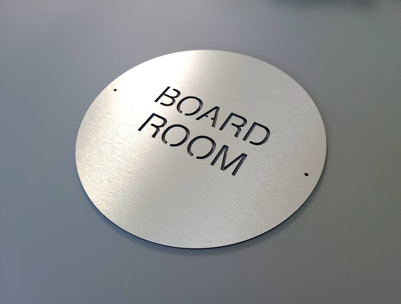 May include: A circular sign made of brushed metal, featuring the cut-out text "BOARD ROOM". The sign is silver with a matte finish. Two small holes are drilled near the top and bottom. The background is grey.
