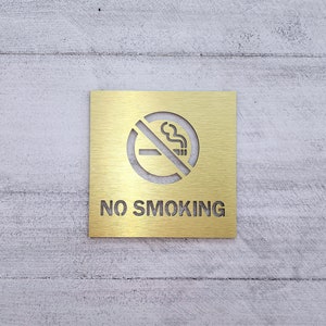 May include: A gold metal sign with a no smoking symbol and the text "NO SMOKING" on a white wood background.