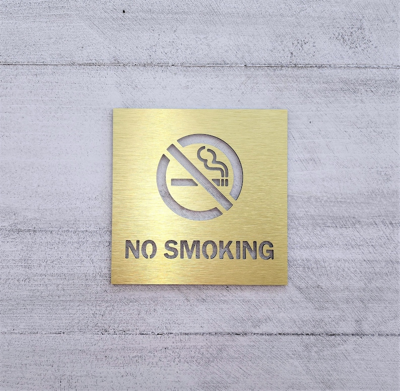May include: A gold metal sign with a no smoking symbol and the text "NO SMOKING" on a white wood background.