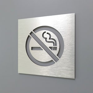 Könnte beinhalten: Ein silbernes Metallschild mit einem Nichtraucher-Symbol. Das Symbol ist ein Kreis mit einem diagonalen Strich durch ihn und einem stilisierten Bild einer Zigarette mit Rauch.