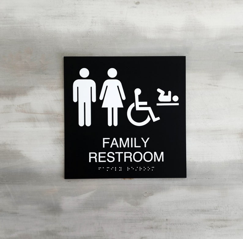 Pu&ograve; includere: Un cartello bianco e nero con la scritta "FAMILY RESTROOM" in lettere bianche. Il cartello include anche icone per uomini, donne, accesso per disabili in sedia a rotelle e un fasciatoio.