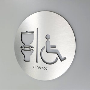 May include: A round, silver-colored sign with a toilet and wheelchair symbol cut out. The sign also includes braille text.