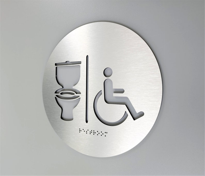 May include: A round, silver-colored sign with a toilet and wheelchair symbol cut out. The sign also includes braille text.