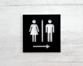 May include: A black square restroom sign with white gender symbols and an arrow pointing right. The sign features a female figure on the left and a male figure on the right, separated by a vertical line.
