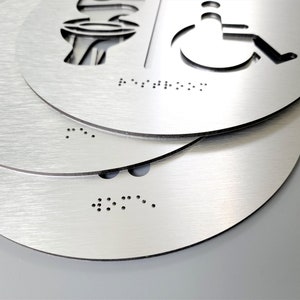 May include: Three silver round signs with braille markings. The top sign has a cut-out image of a wheelchair and toilet. The bottom two signs have braille markings only.