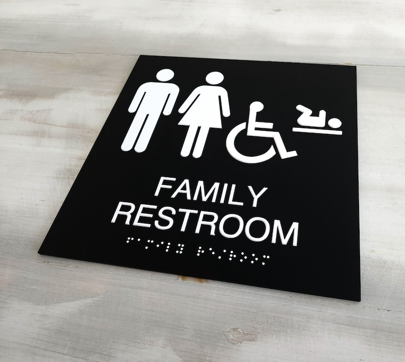 Pu&ograve; includere: Segnale bianco e nero con il testo "FAMILY RESTROOM" in lettere bianche. Il segnale include anche icone per uomini, donne, accesso per disabili in sedia a rotelle e fasciatoio.