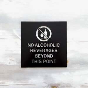 May include: Black metal sign with a white silhouette of a wine bottle and glass with a diagonal line through it. The sign reads "No Alcoholic Beverages Beyond This Point."