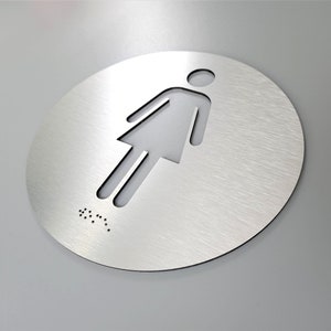 May include: A round, silver-colored metal sign with a cutout of a female figure. The sign has braille lettering at the bottom.