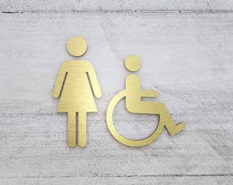 Könnte beinhalten: Goldfarbene Metallschildchen für Toiletten an einer weißen, strukturierten Wand. Die Schilder zeigen eine weibliche Figur und eine Person im Rollstuhl.