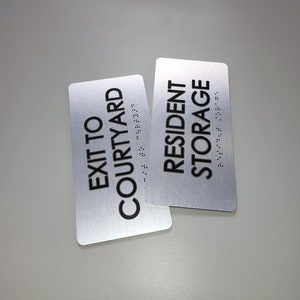 K&ouml;nnte beinhalten: Zwei silberne Schilder mit schwarzem Text. Das obere Schild lautet "Resident Storage" und das untere Schild lautet "Exit to Courtyard". Beide Schilder haben Braille-Schrift.