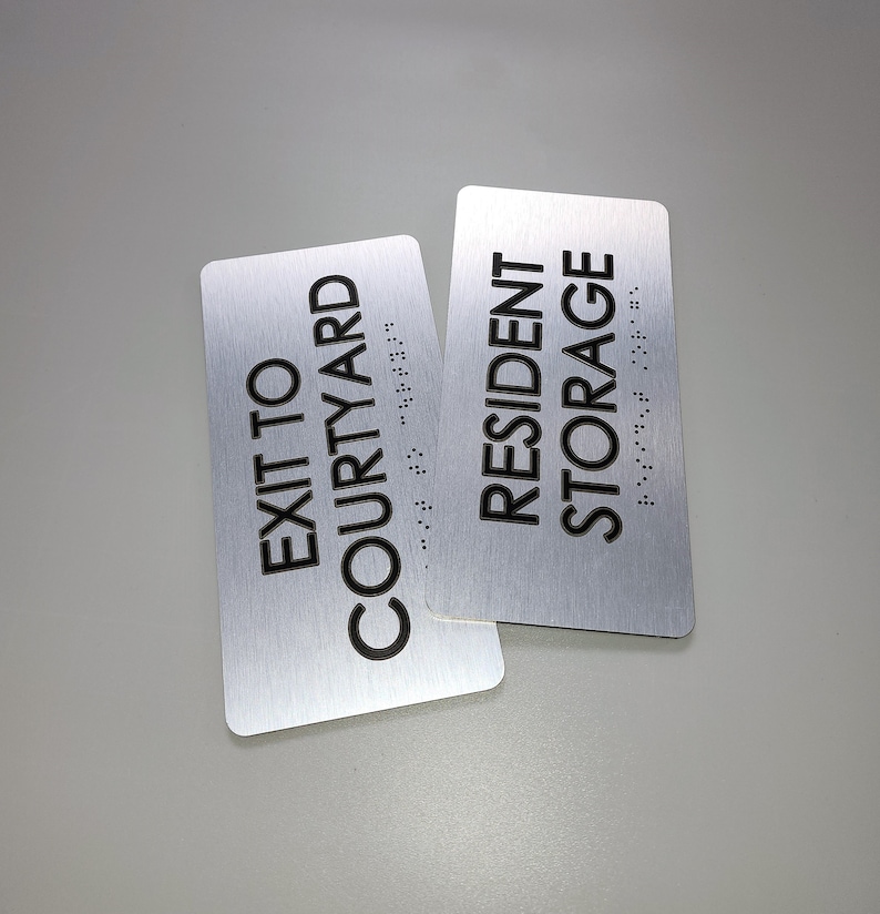 K&ouml;nnte beinhalten: Zwei silberne Schilder mit schwarzem Text. Das obere Schild lautet "Resident Storage" und das untere Schild lautet "Exit to Courtyard". Beide Schilder haben Braille-Schrift.