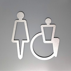 Pode incluir: Placa de metal prateada com um símbolo feminino e um símbolo de pessoa em cadeira de rodas, indicando um banheiro para ambos os sexos.